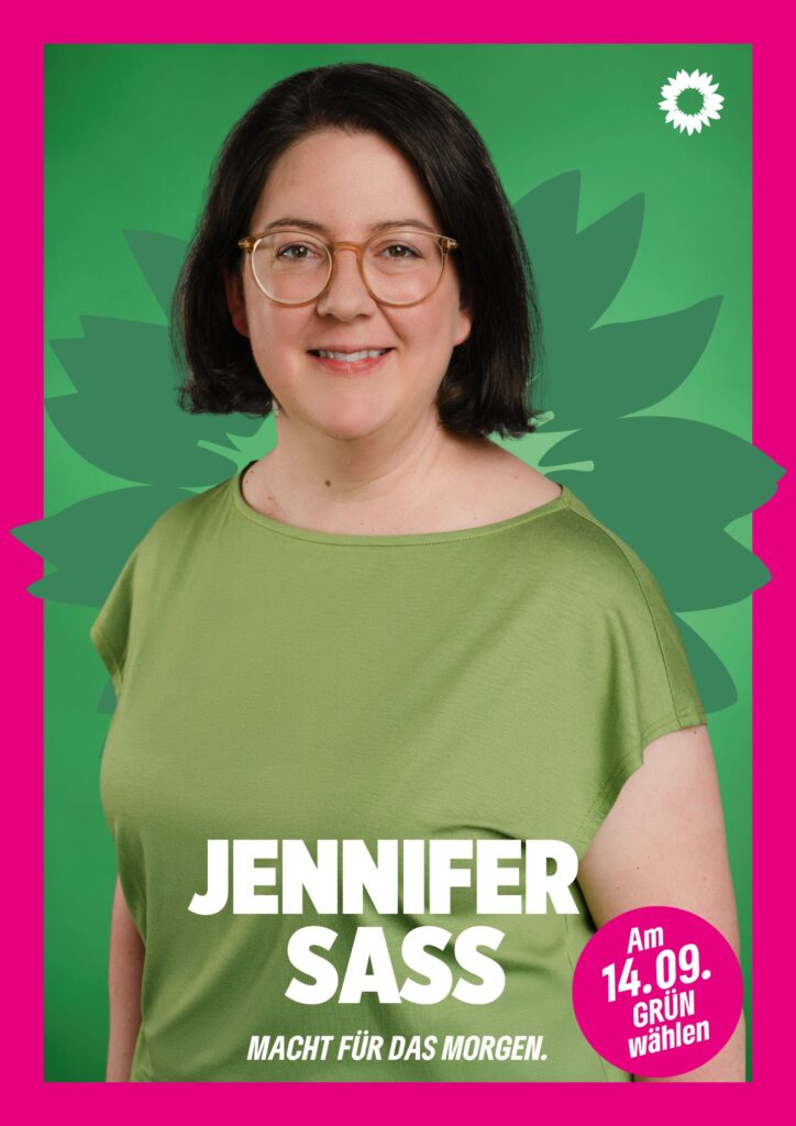 Portraitaufnahme von Jennifer Sass als Wahlplakat mit grüner Sonnenblume im Hintergrund, weißer kleiner Sonnenblume oben rechts, seinem Namen, einem Button mit dem Text "Am 14.09. Grün wählen" und dem Claim Macht für das Morgen.