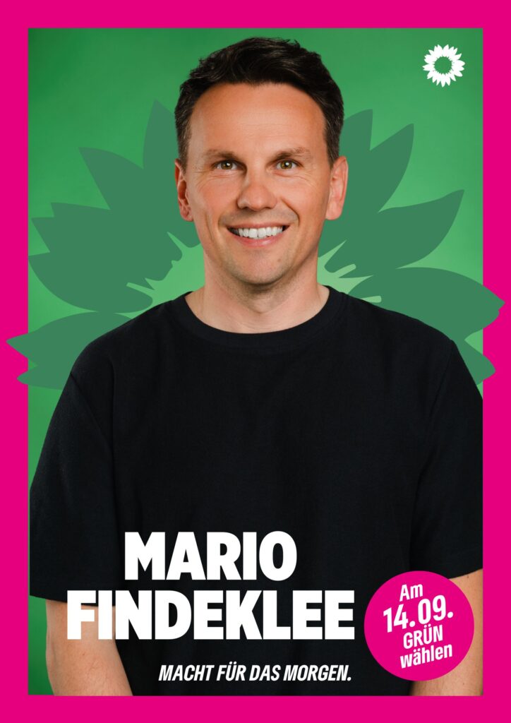 Portraitaufnahme von Mario Findeklee als Wahlplakat mit grüner Sonnenblume im Hintergrund, weißer kleiner Sonnenblume oben rechts, seinem Namen, einem Button mit dem Text "Am 14.09. Grün wählen" und dem Claim Macht für das Morgen.
